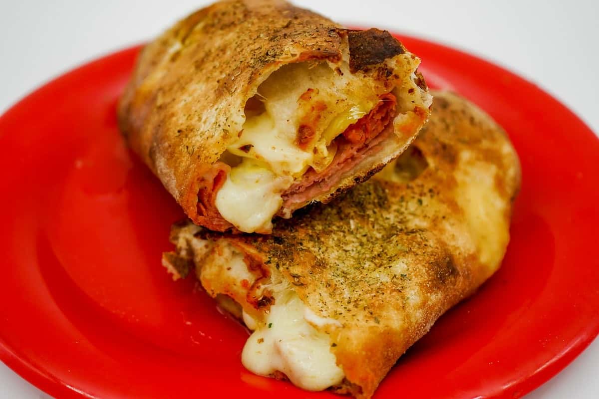 Golden-brown calzone stuffed with melted cheese, meats, and peppers, served on a vibrant red plate.
