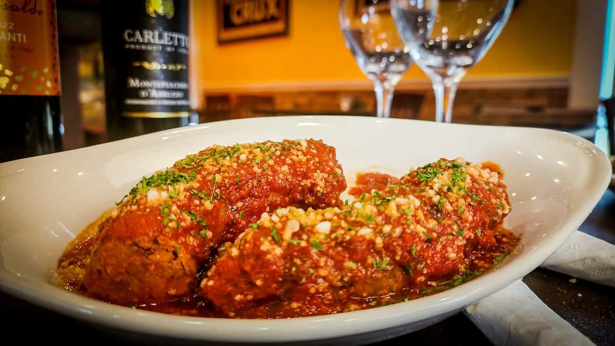 Italian-style meatballs in marinara sauce, topped with Parmesan and parsley, served in a white bowl with wine bottles and glasses behind.
