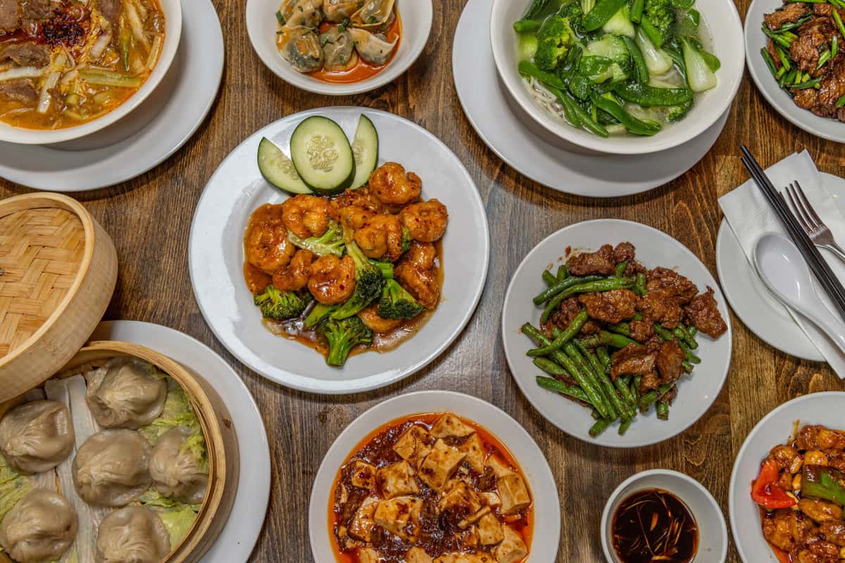 Assorted Chinese stir-fry dishes served on white plates, including spicy chicken with chili sauce, shrimp and broccoli in garlic brown sauce, Kung Pao chicken with bell peppers and peanuts, beef with scallions, and beef with green beans, all presented on a rustic wooden table.