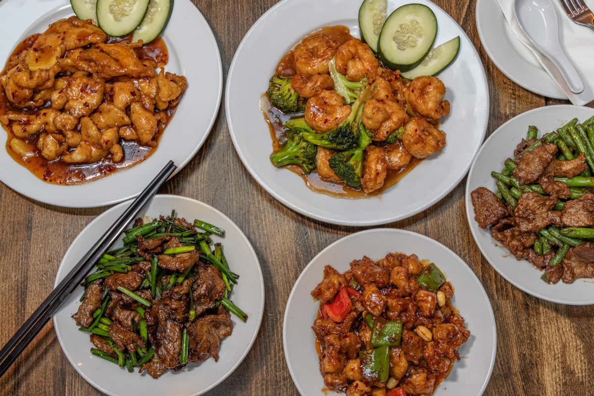 Assorted Chinese stir-fry dishes served on white plates, including spicy chicken with chili sauce, shrimp and broccoli in garlic brown sauce, Kung Pao chicken with bell peppers and peanuts, beef with scallions, and beef with green beans, all presented on a rustic wooden table.