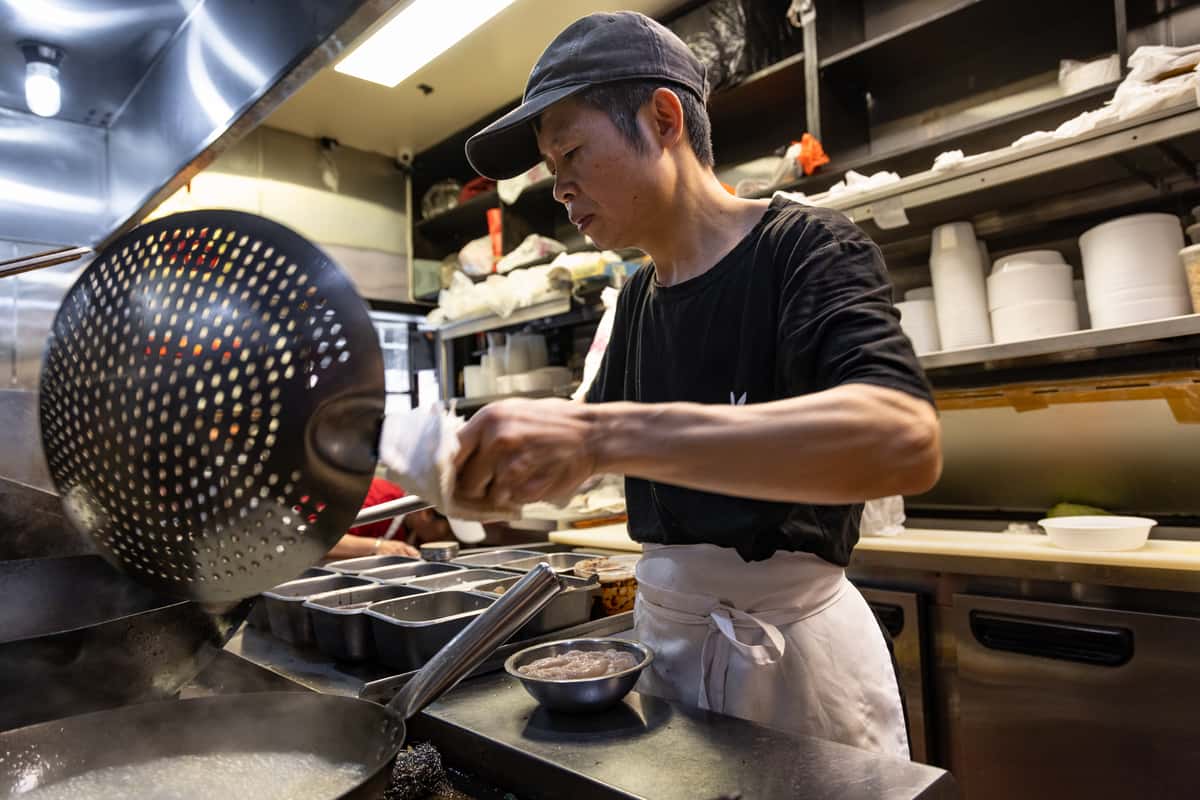 Chinese chef preparing noodle soup in a bustling restaurant kitchen, using a perforated ladle to drain ingredients over a steaming wok, with fresh ingredients and stainless steel equipment in the background.
