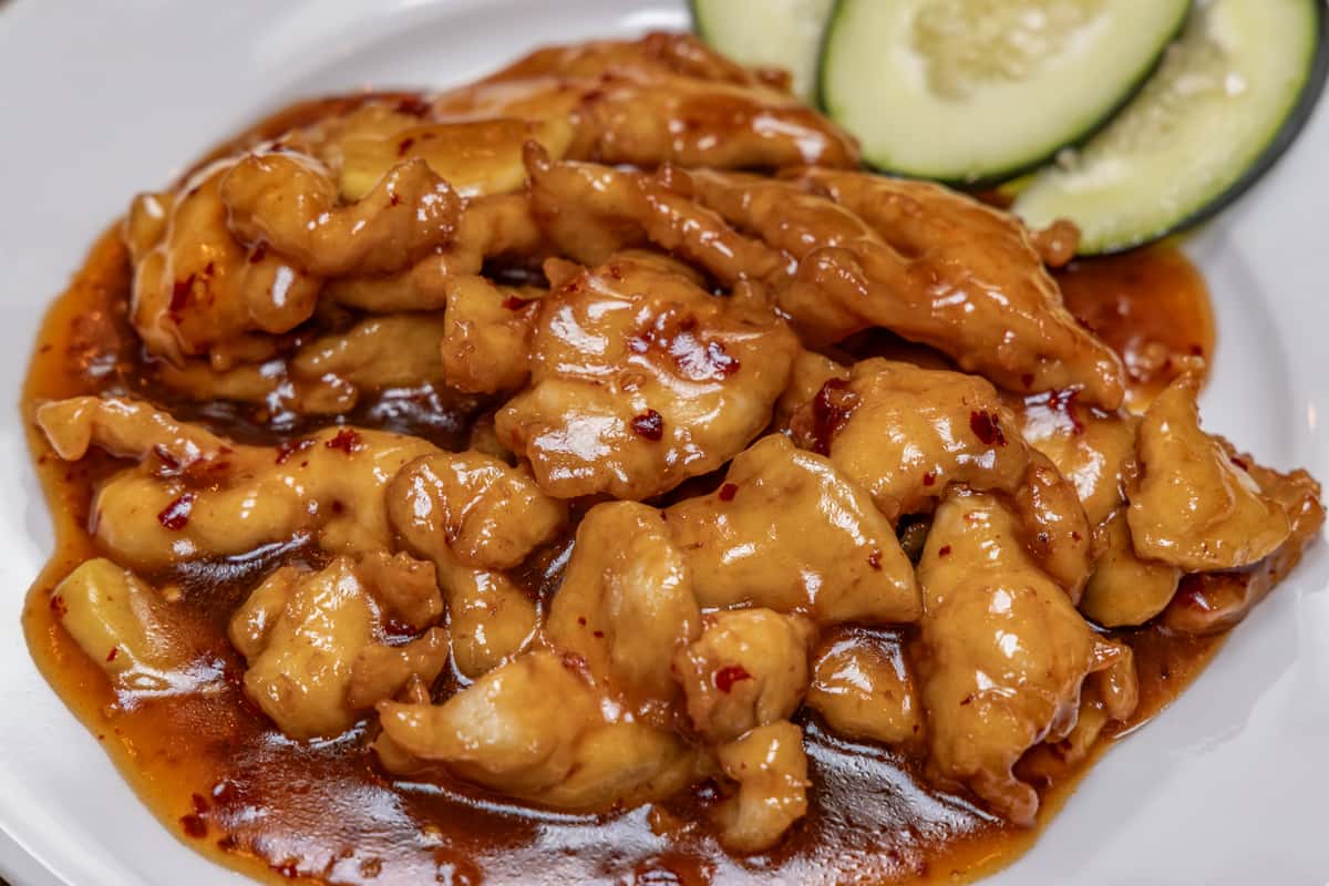 Close-up of spicy Szechuan-style chicken in a glossy chili garlic sauce, garnished with cucumber slices on the side, served on a white plate at a Chinese restaurant.
