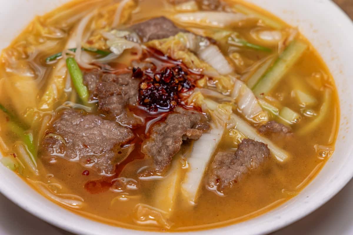 Bowl of spicy beef noodle soup with tender slices of beef, napa cabbage, scallions, and chili oil in a rich, savory broth, served in a white bowl at a Chinese restaurant.