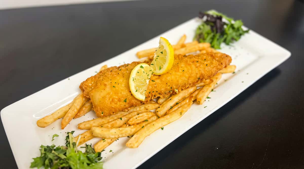 Fried fish fillet and french fries with lemon slices and greens on a white platter.