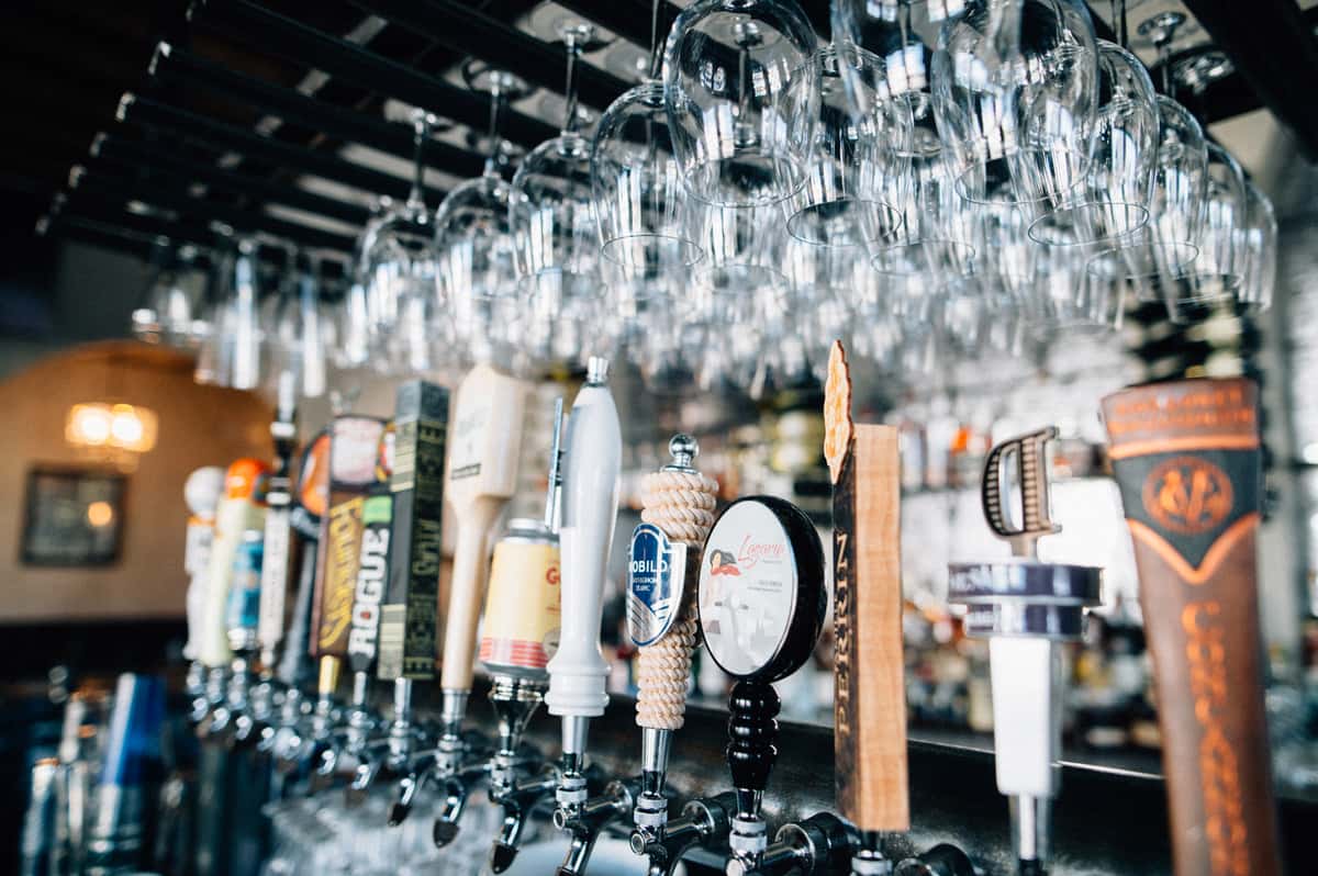 Variety of beers on tap with wine glasses hanging above on a wine rack.