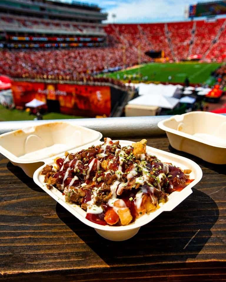 Takeout tray of loaded fries held in front of a stadium with a red seating section and green field