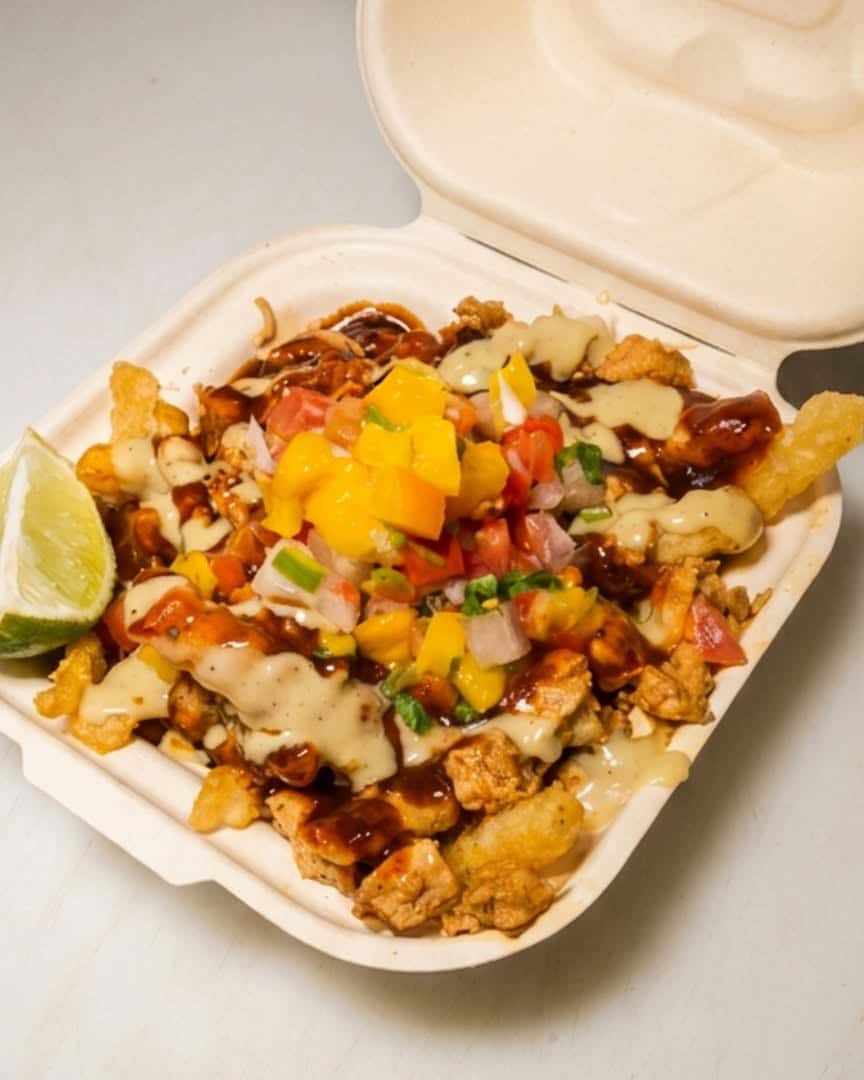 Loaded fries topped with sauce, cheese, and garnish served in a takeout container, with a lime wedge on the side