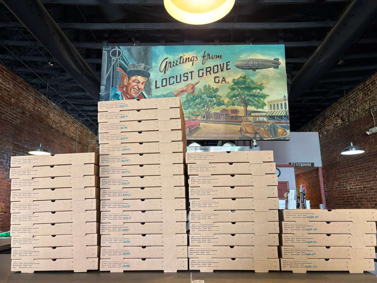 Stacks of pizza boxes inside a rustic Locust Grove, Georgia restaurant beneath a vintage "Greetings from Locust Grove GA" mural.