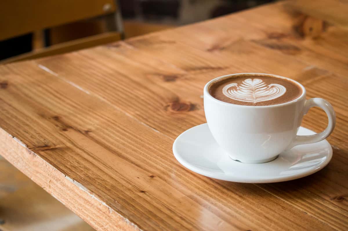 A beautifully crafted cup of latte with intricate leaf-shaped latte art, served in a classic white ceramic cup and saucer, set on a rustic wooden table—evoking a cozy café atmosphere.