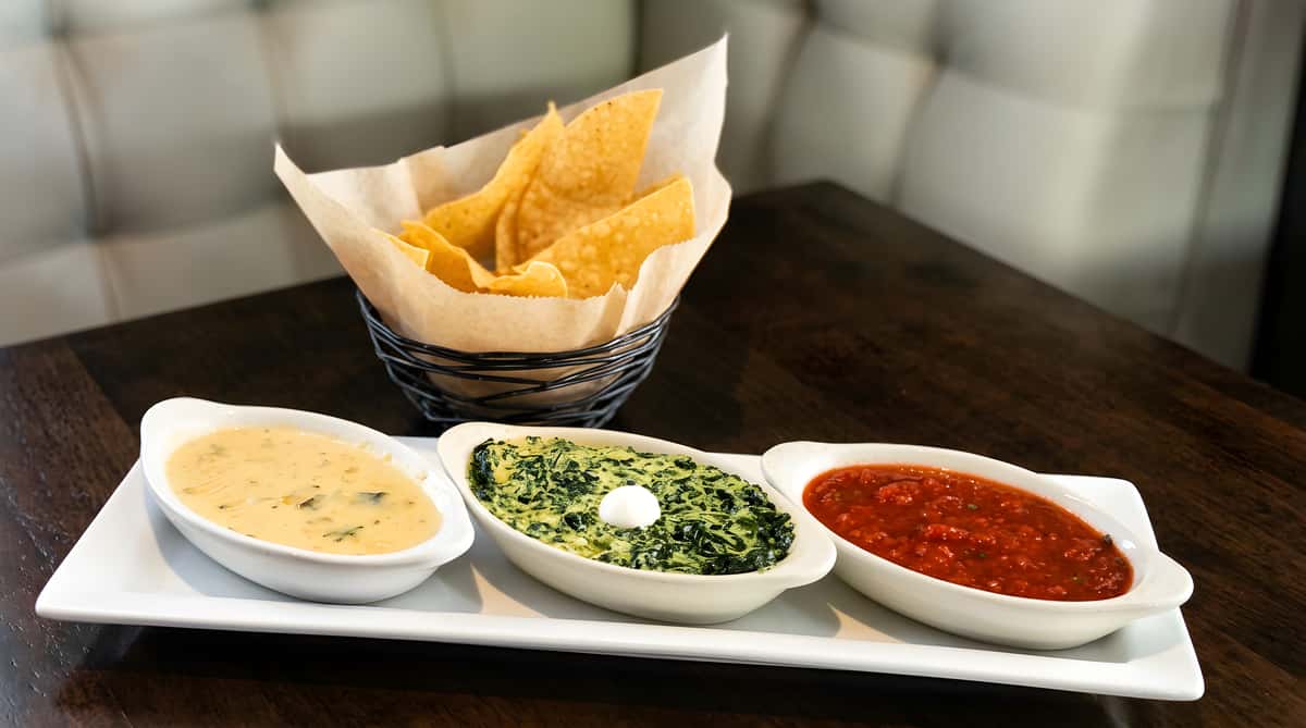 Chips and dips platter with queso, spinach artichoke dip, and red salsa on a white tray.