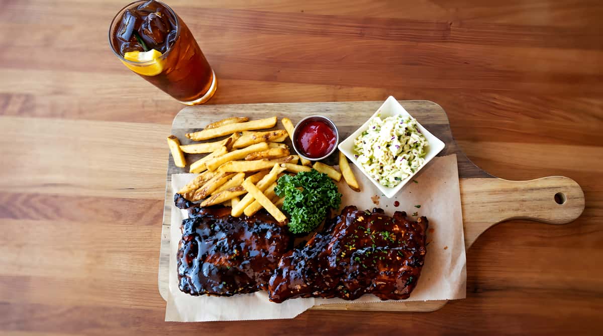 BBQ ribs with fries, a side of coleslaw, ketchup, and a glass of iced tea on a wooden board.