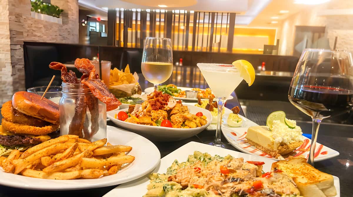 A table filled with various dishes: a burger with fries, pasta with grilled chicken, salad, cheesecake, and drinks in a restaurant with blue lighting.