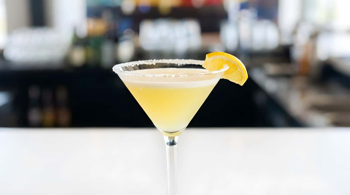 Martini glass with a pale yellow drink, a sugar-rimmed edge, and a lemon slice garnish.