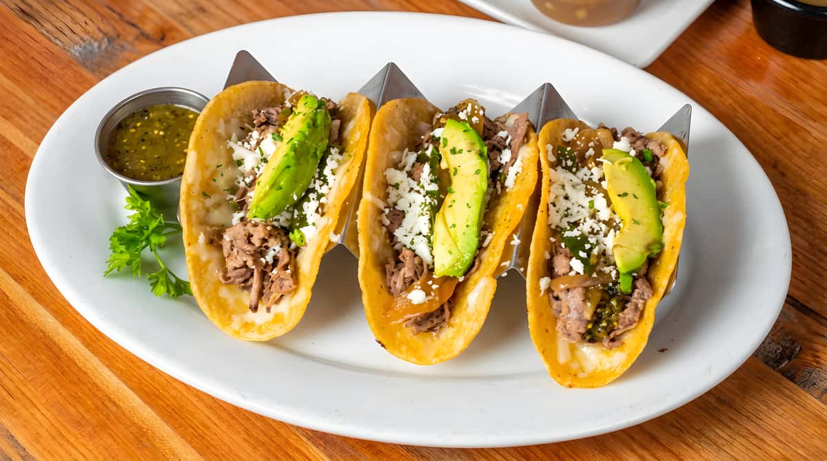 Four street tacos filled with meat, crumbled white cheese, and avocado slices, served with green salsa.