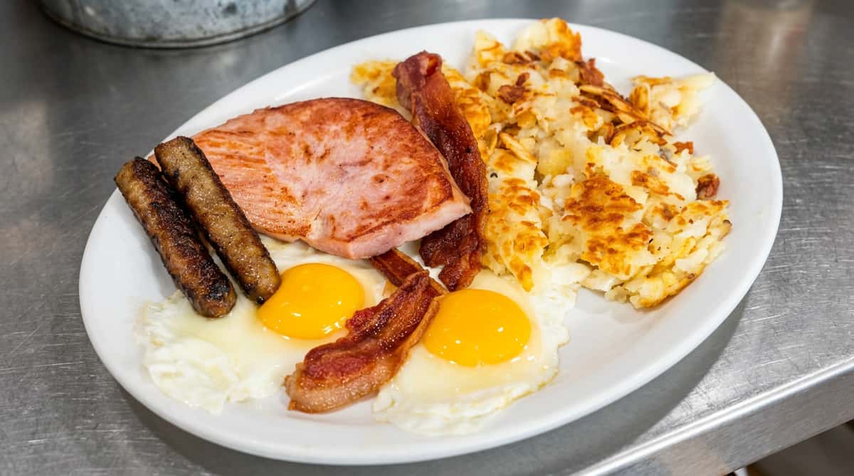 Breakfast plate with hash browns, two fried eggs, sausage links, thick-cut bacon, and a slice of ham.