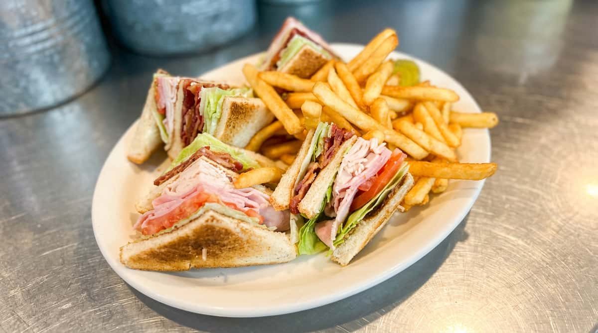 Club sandwich with turkey, ham, bacon, lettuce, tomato, and fries on a white plate with a pickle.