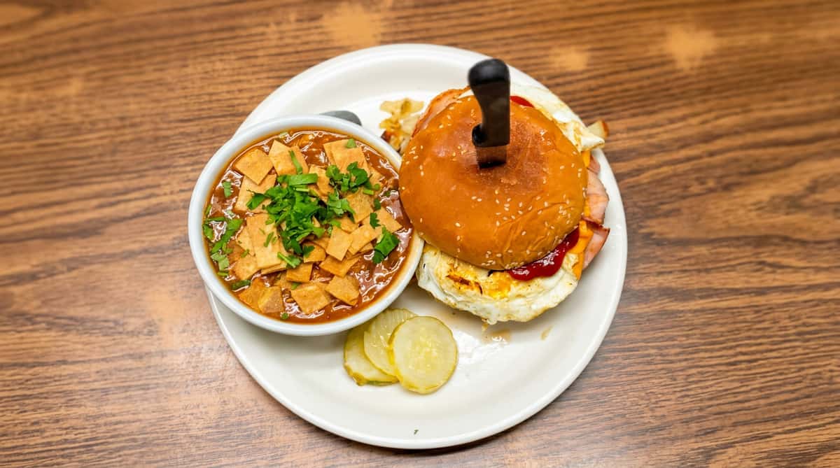 Hamburger with fried egg, cheese, and bacon, skewered with a knife, next to a bowl of soup with tortilla strips and cilantro, and sliced pickles on a white plate.