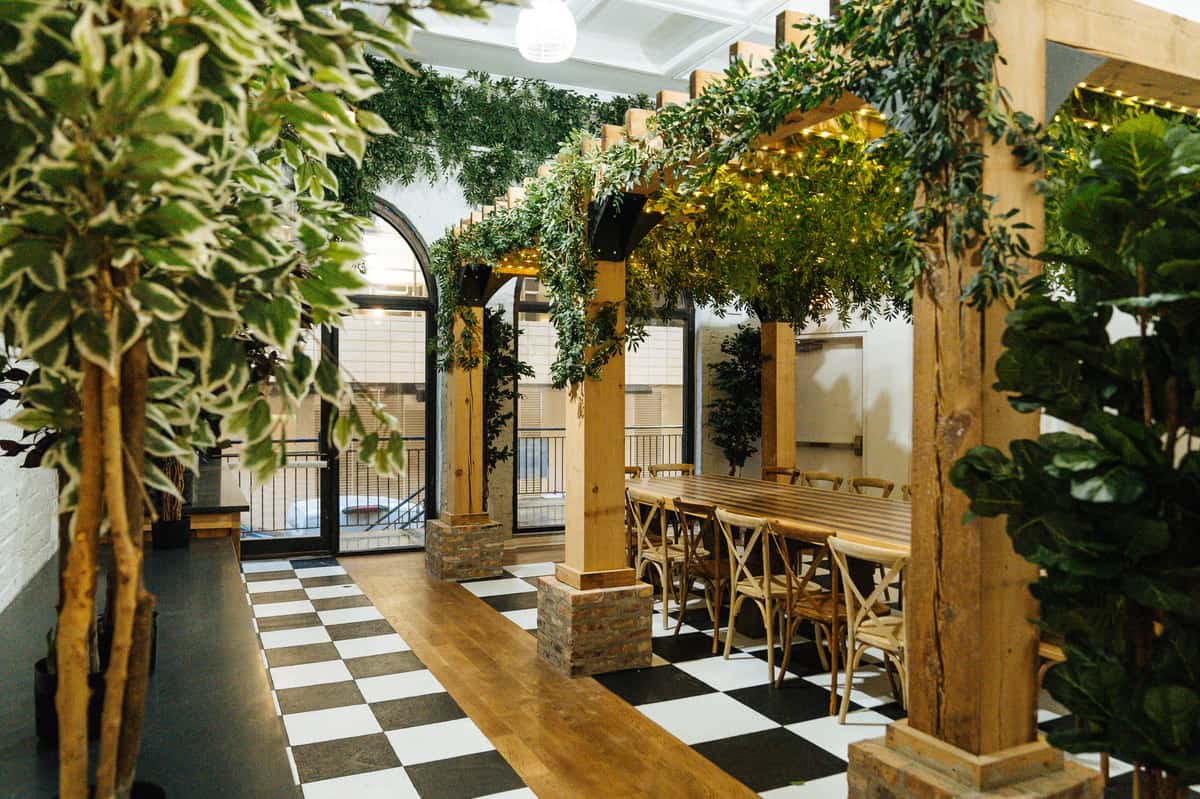 Elegant indoor dining space with a wood pergola draped in greenery, checkerboard and wood flooring, and natural light.