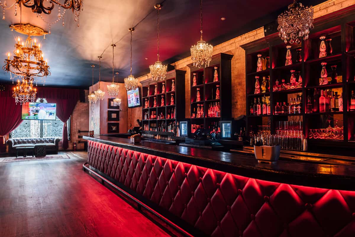 Luxurious bar with a red tufted front, backlit liquor shelves, chandeliers, and rich red drapery creating a warm, elegant atmosphere.