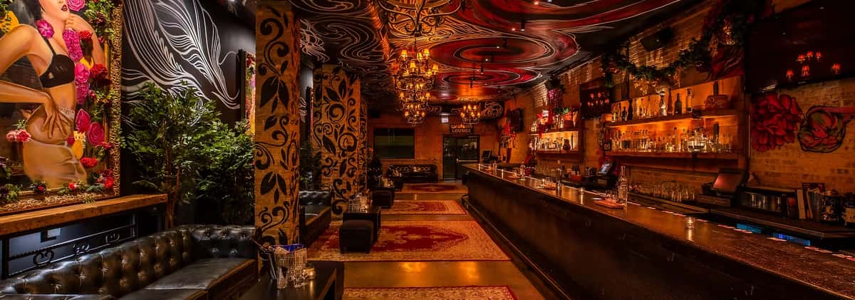 Lounge bar with ornate murals, chandeliers, black leather seating, and a long bar under a swirled, painted ceiling.