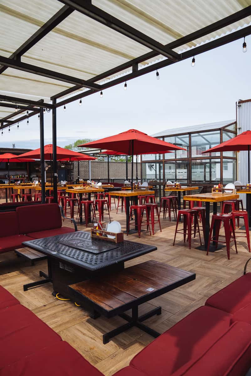 Outdoor rooftop dining space with red umbrellas, high-top tables, and wooden floors under a partially covered pergola.