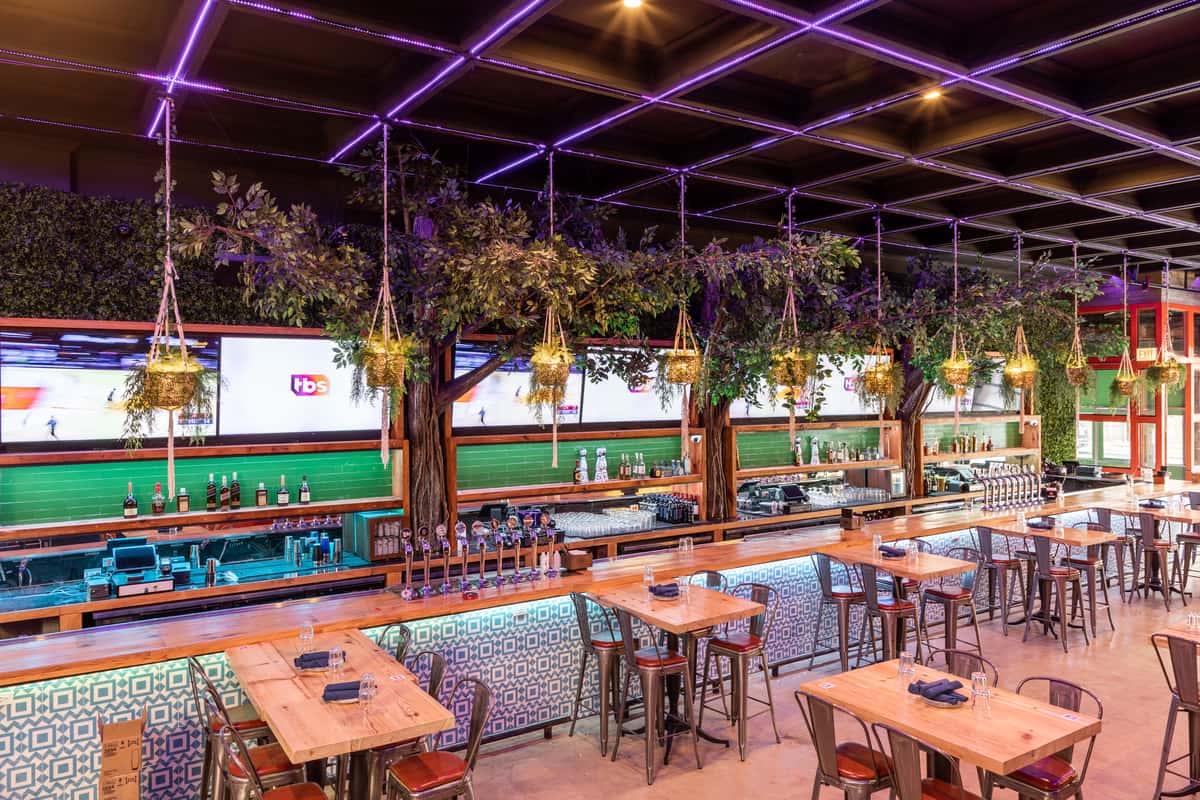 A stylish bar and restaurant with wooden tables, hanging plants, neon ceiling lights, and large TVs above a fully stocked bar.