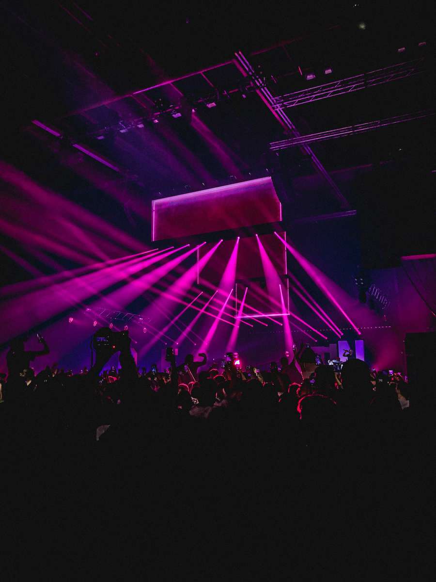Crowd at a concert under dramatic magenta stage lights with beams cutting through a dark venue ceiling.