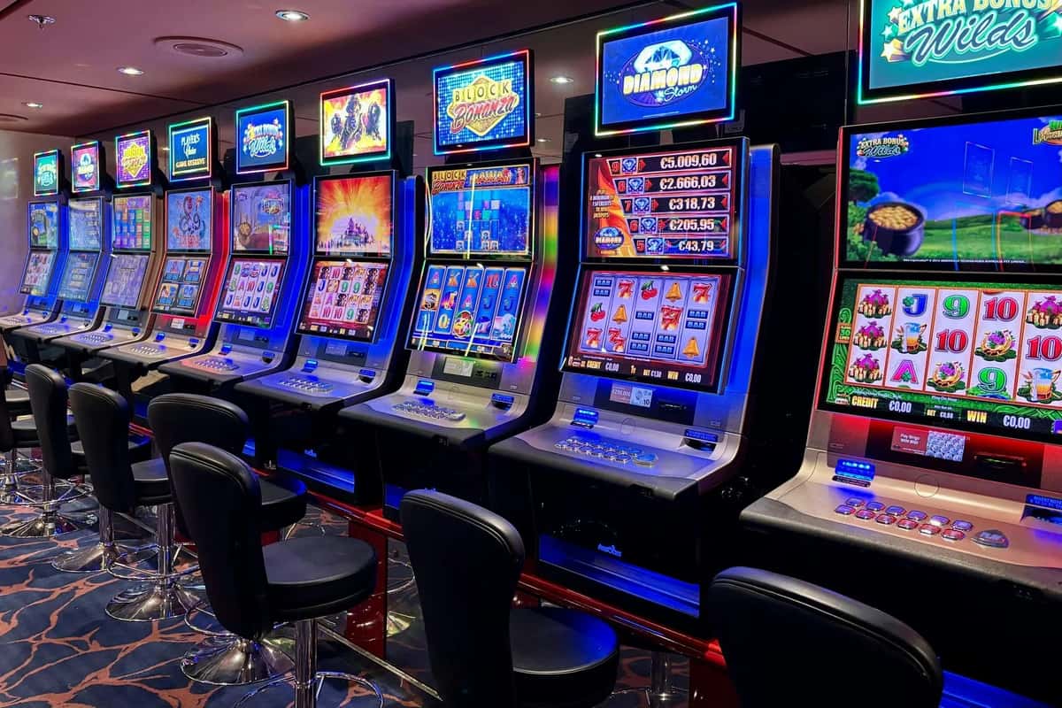 Row of colorful slot machines with flashing screens in a modern casino, empty chairs lined up in front.