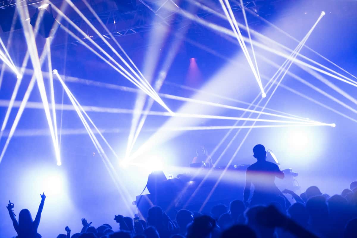 Crowd dancing under intense white and blue laser lights at a nightclub concert with a DJ performing on stage.