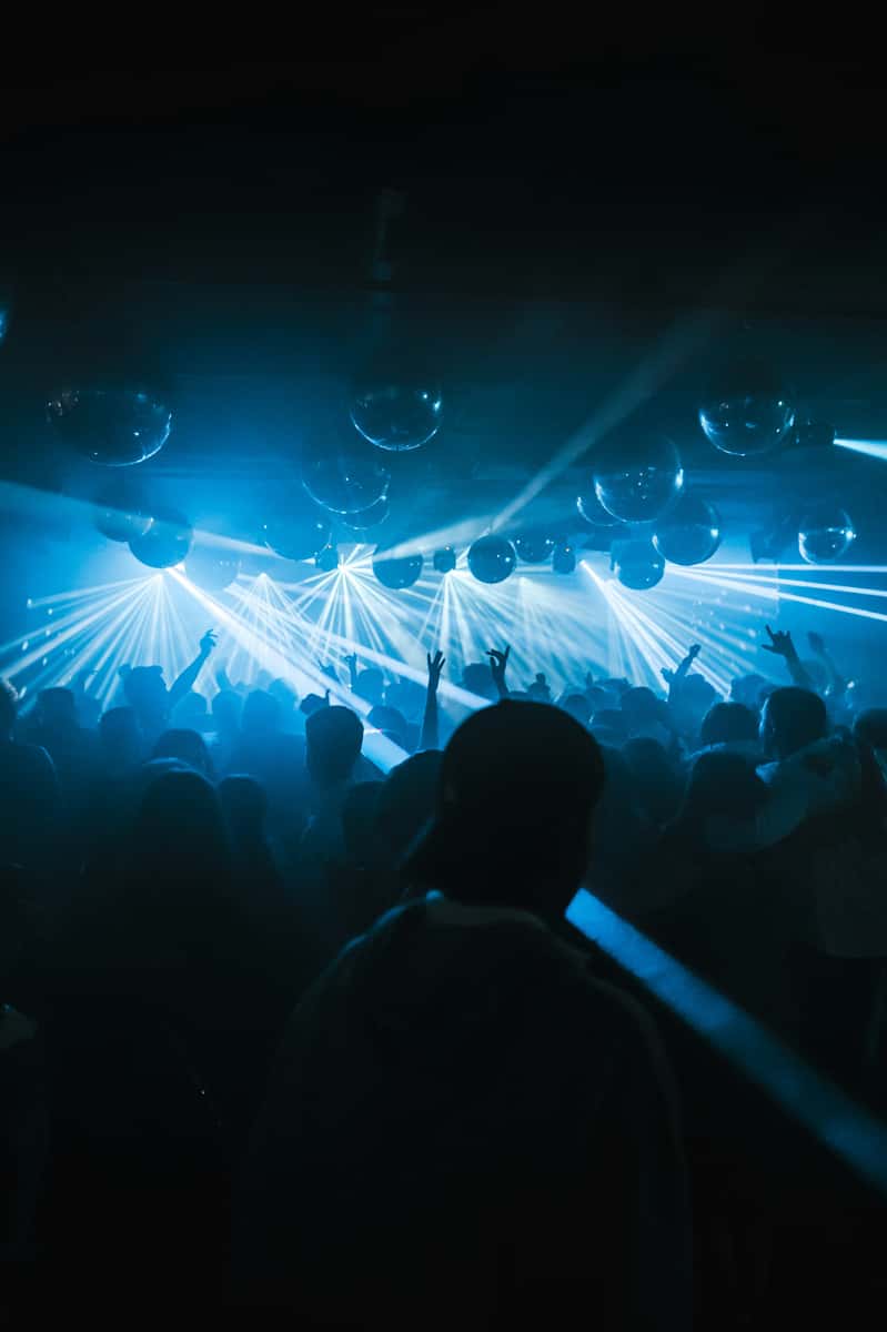Crowd dancing under blue lights and disco balls in a packed nightclub with beams of light cutting through the haze.