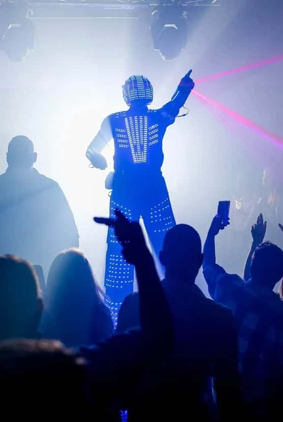 Performer in a glowing LED suit on stage with laser lights, surrounded by a cheering nightclub crowd.