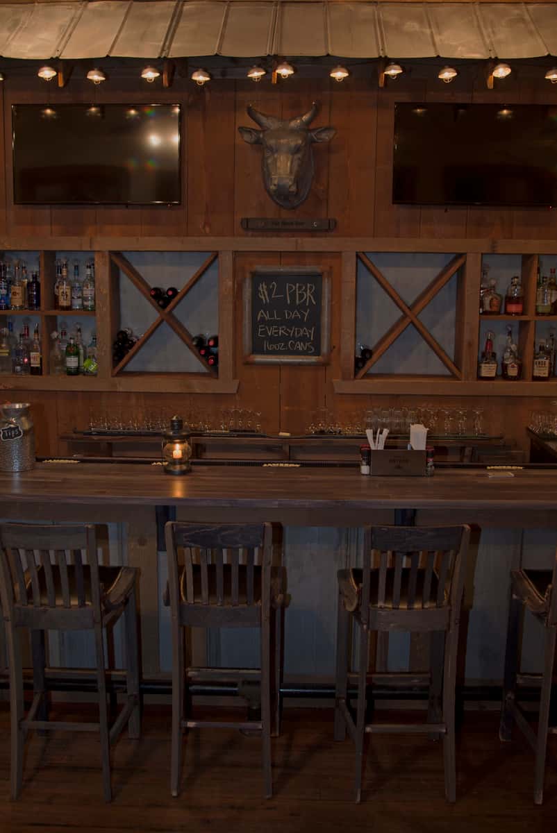 A rustic bar features wooden chairs, shelves of alcohol, and a mounted bull head. A chalkboard sign advertises "$2 PBR ALL DAY EVERYDAY TALL CANS".