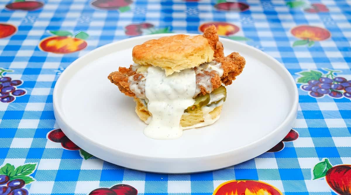 Fried chicken biscuit with pickles and a drizzle of creamy white sauce, on a white plate.