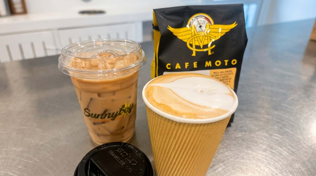 Iced coffee in a plastic cup, a latte with art in a paper cup, and a bag of Cafe Moto coffee beans.