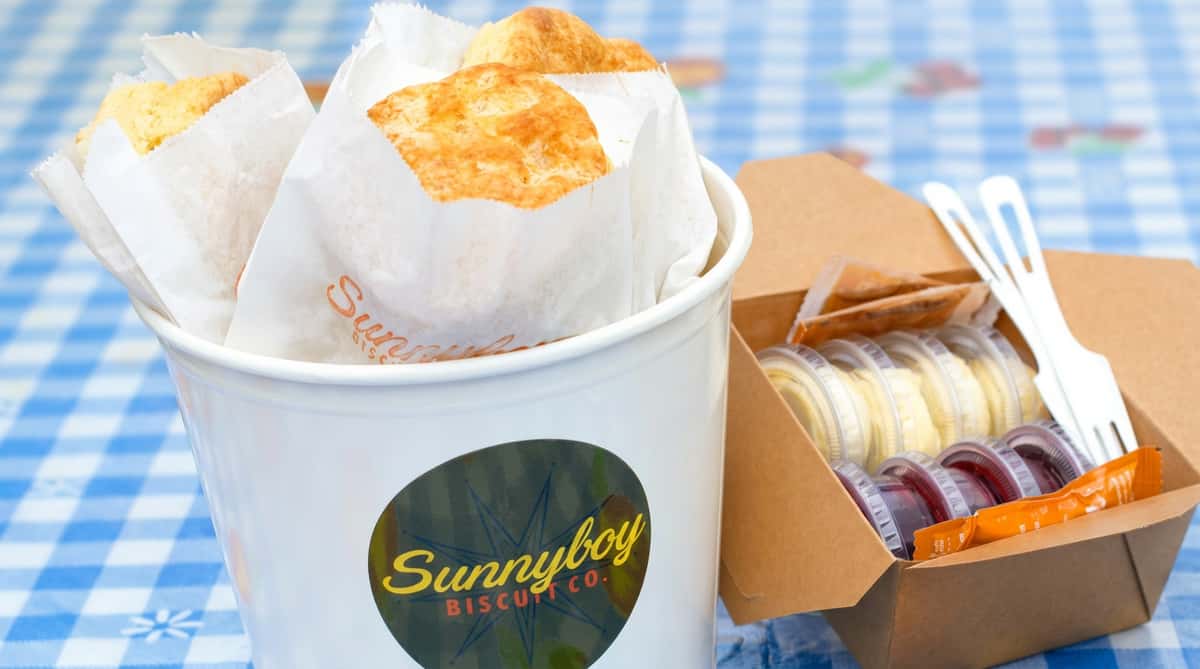 Biscuits in paper sleeves in a "Sunnyboy Biscuit Co." bucket, with a takeout box of sauces and plastic cutlery next to it.