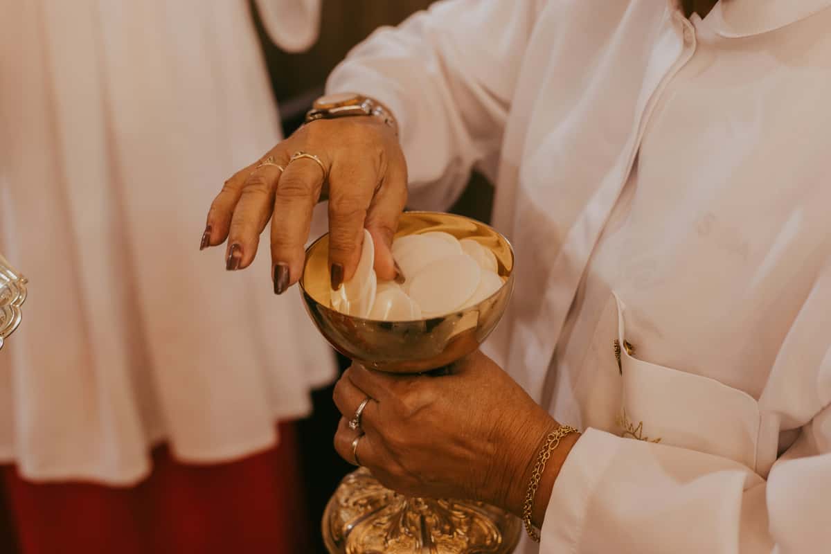 Person in white religious attire holding a golden chalice filled with communion wafers during a church service.