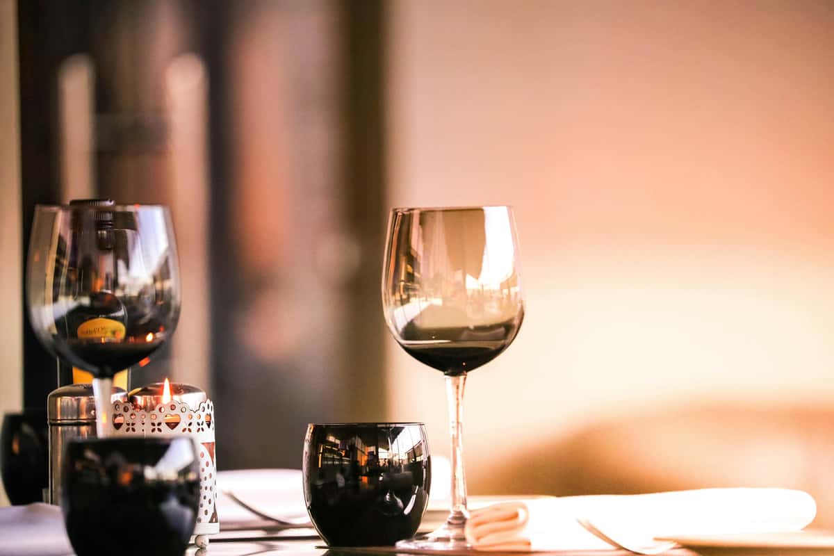 Two glasses of red wine and a smaller wine glass sit on a softly lit table beside a candle holder, creating a warm, intimate dining atmosphere.