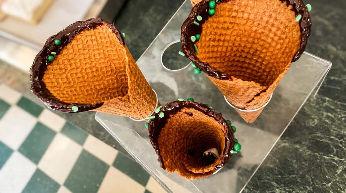 Three waffle cones with chocolate dipped rims and green sprinkles, sitting in a clear acrylic stand on a green marble counter.