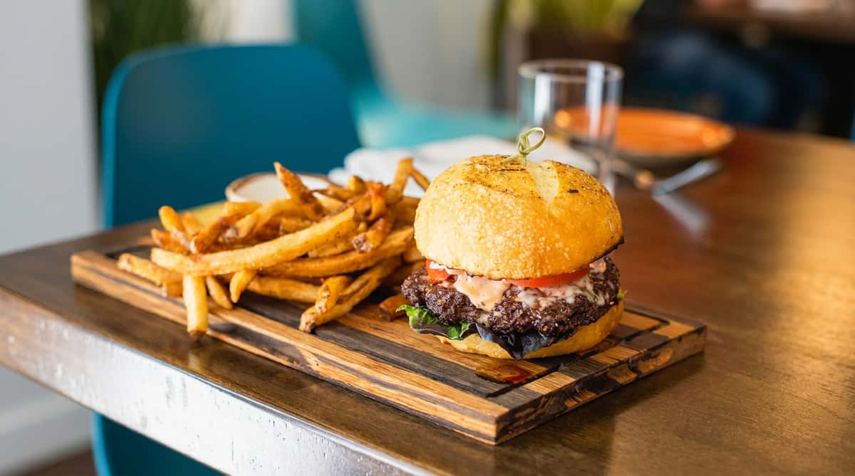 Cheeseburger with a toasted bun, melted cheese, tomato, lettuce, and fries on a wooden board.