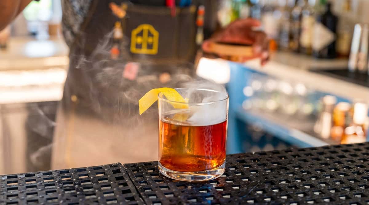 Cocktail in a short glass with amber liquid, lemon peel garnish, and smoke rising from the top.