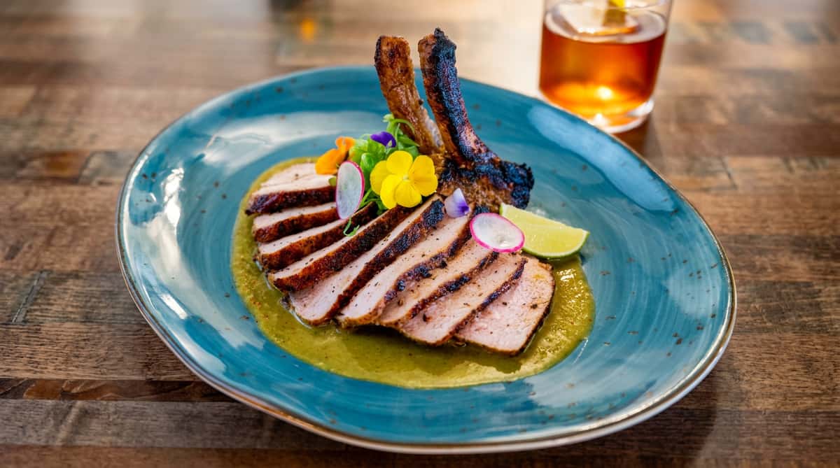 Sliced pork chop with bones on a bed of green sauce, garnished with edible flowers, radish slices, and lime.