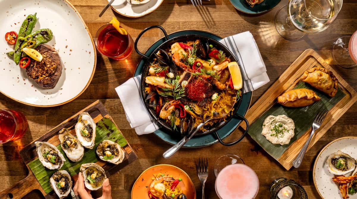 Seafood paella with shrimp, mussels, and fish in a pan, surrounded by oysters, empanadas, and cocktails on a wooden table.