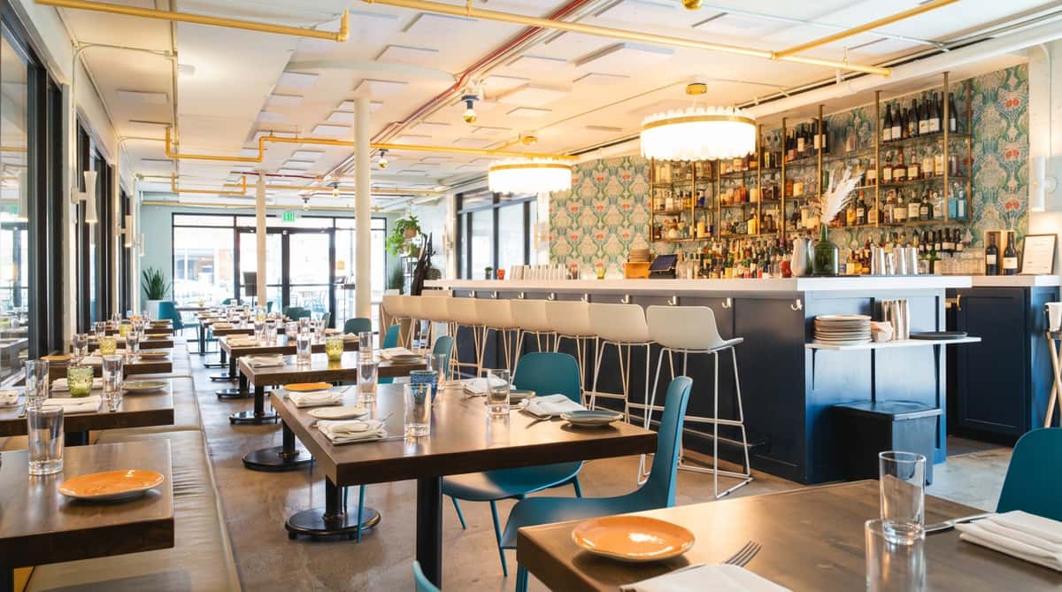 Restaurant dining room with wooden tables, teal chairs, a bar with white stools, and patterned wallpaper.