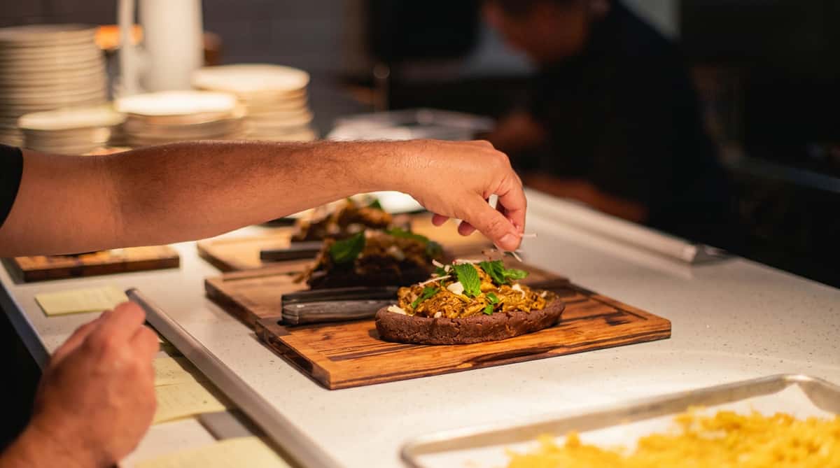 Chef garnishing a savory tart or flatbread with shredded white garnish and mint leaves on a wooden board.