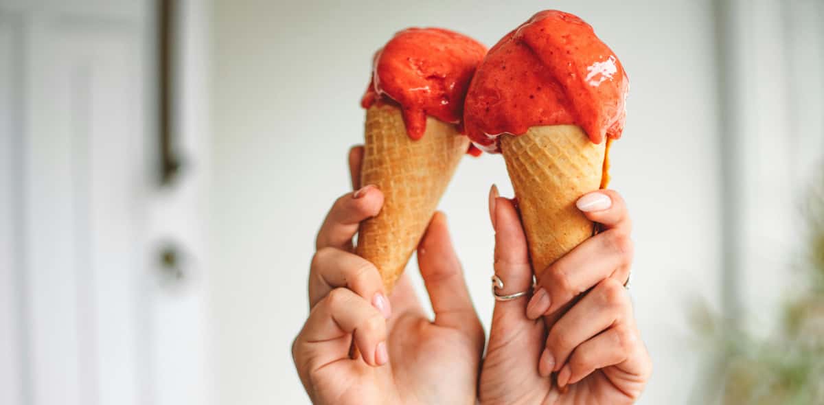 Two hands holding strawberry ice cream cones with scoops slightly melting.