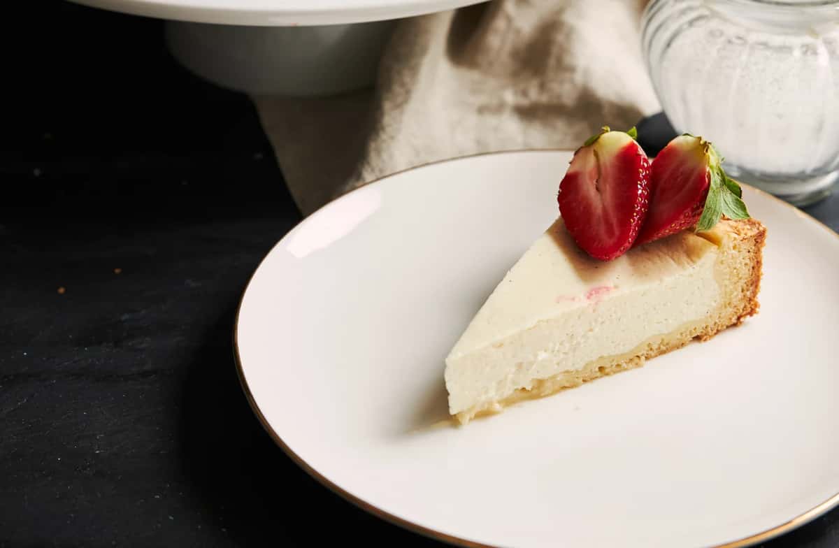 Slice of classic New York–style cheesecake topped with fresh strawberries on a white plate, highlighting a creamy texture and elegant café dessert presentation.