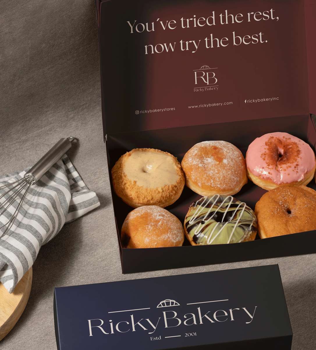 Open box of assorted gourmet donuts from Ricky Bakery, featuring powdered sugar, glazed, and chocolate-drizzled varieties, styled in branded packaging with elegant bakery branding for a premium dessert gift presentation.