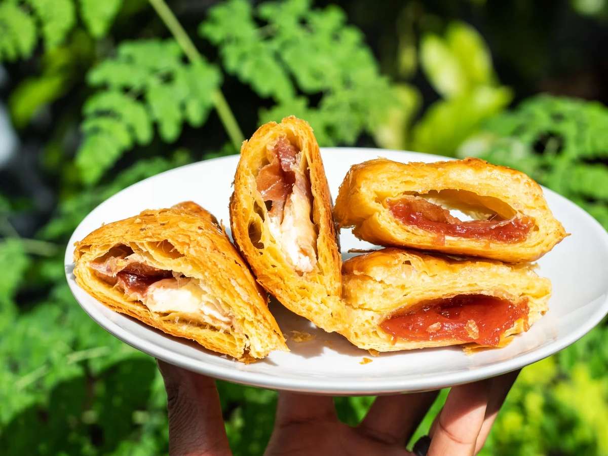 Flaky puff pastry turnovers filled with melted cheese and cured meat alongside a sweet fruit-filled pastry, served on a white plate outdoors, highlighting golden layers and a fresh café-style brunch presentation.