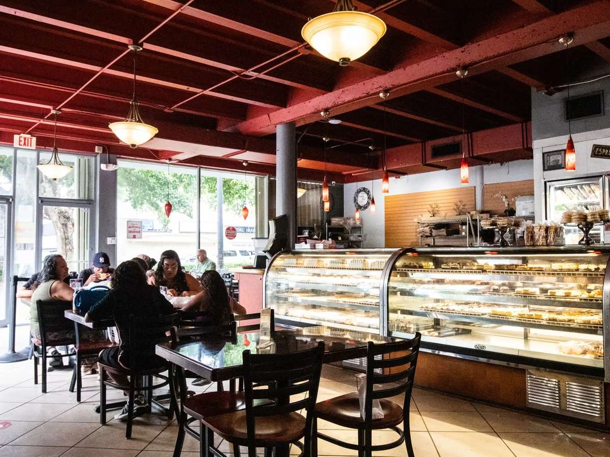 Cozy neighborhood café interior with customers dining at tables beside a glass pastry display case filled with fresh baked goods, warm pendant lighting, and large front windows creating an inviting bakery ambiance.