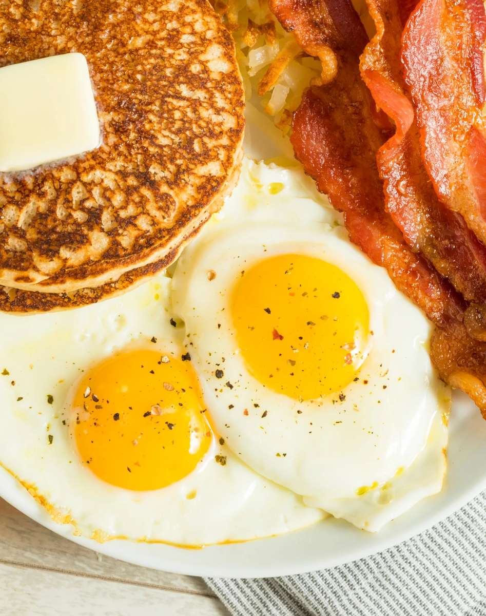 Classic American breakfast plate with fluffy buttermilk pancakes topped with melting butter, crispy bacon strips, golden hash browns, and two sunny-side-up eggs seasoned with cracked black pepper, served fresh in a cozy diner setting.